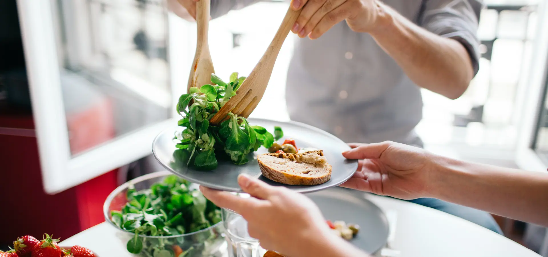 Salat wird mit Hilfe von Holzbesteck auf einen Teller gelegt