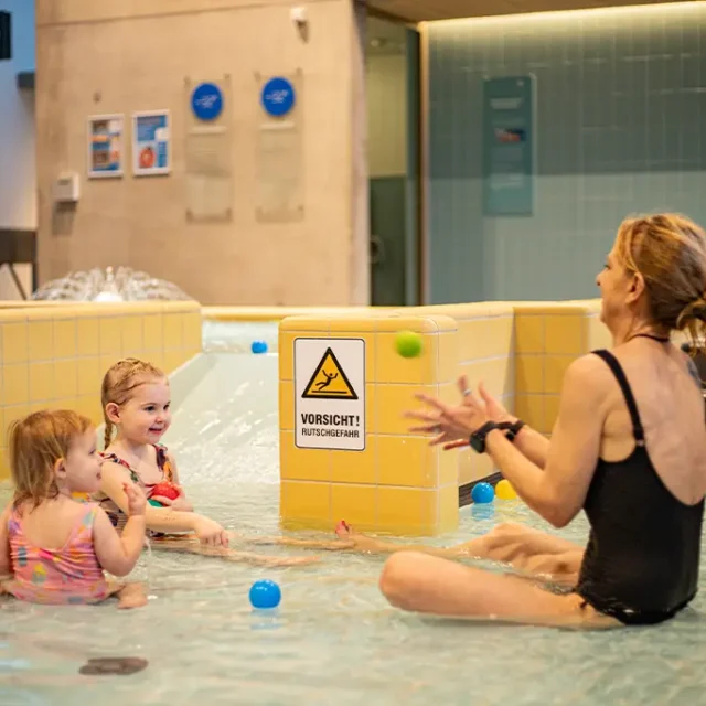 Mama sitzt mit ihren 2 Kindern im Familienbecken und spielt mit bunten Bällen.