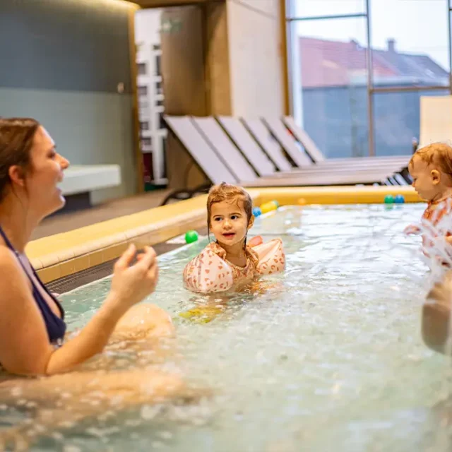 Eltern sitzen mit ihren Kindern im Kleinkinderbecken neben dem Wasserigel