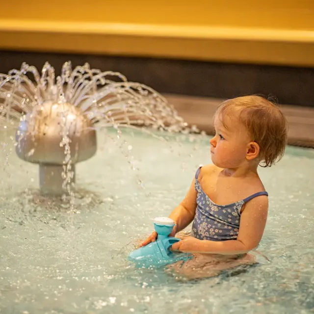 Kind sitzt mit Gießkanne im Kleinkinderbecken neben einem laufenden Wasserigel