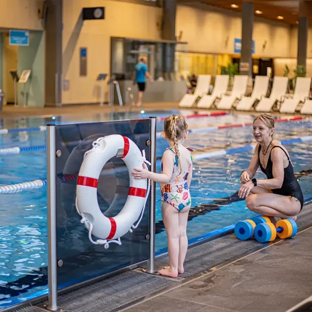 Mama steht mit Kind seitlich vom Sportbecken. Das Kleinkind berührt den Rettungsring.