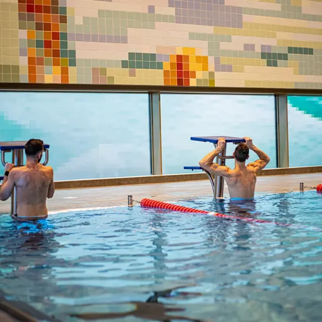 2 Männer ziehen sich im Sportbecken vom Wasser am Startsockel hoch