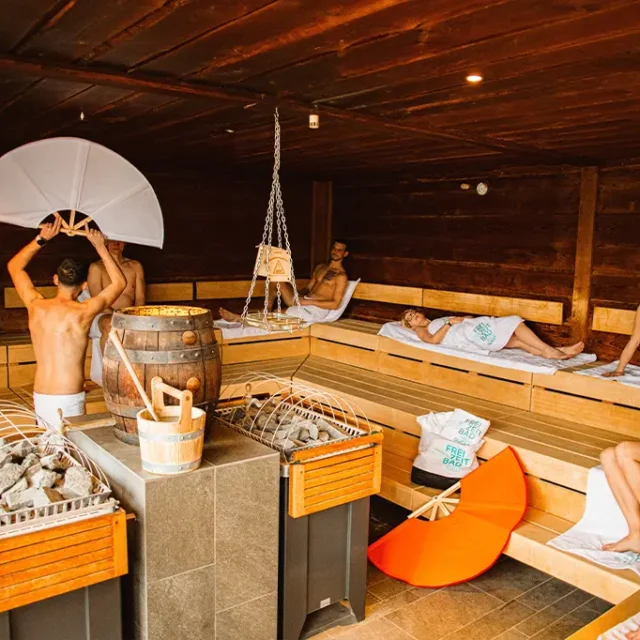 Mann wedelt mit weißem Fächer beim Aufguss in einer Holzsauna mit vielen Gästen