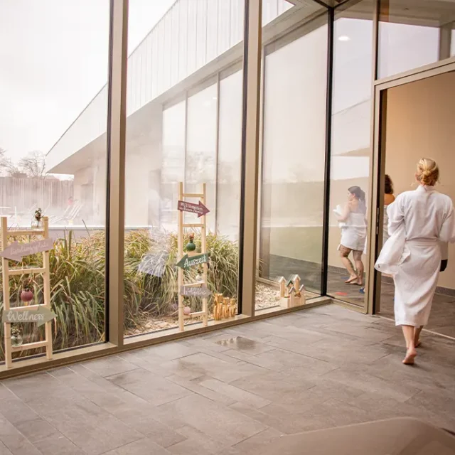 Badegäste in Bademantel laufen aus dem Raum. Blick nach draußen durch die Glasfront, davor zwei Holzleitern mit Dekopfeilen.