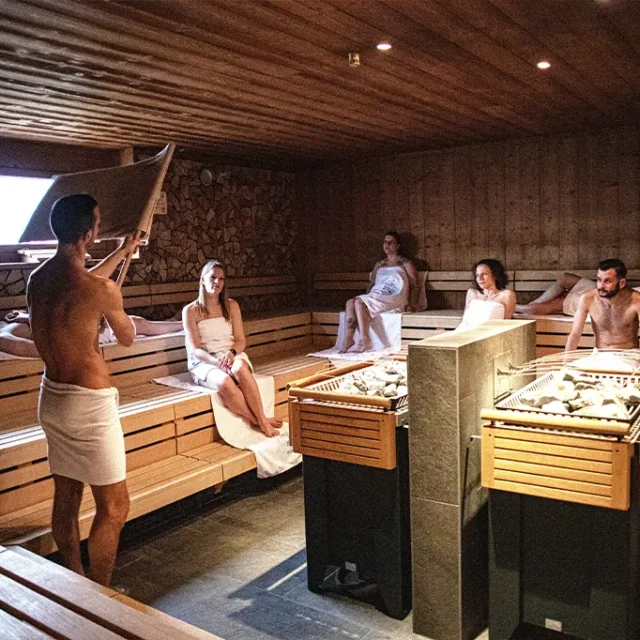 Mann wedelt mit Handtuch beim Aufguss in einer Holzsauna mit vielen Gästen