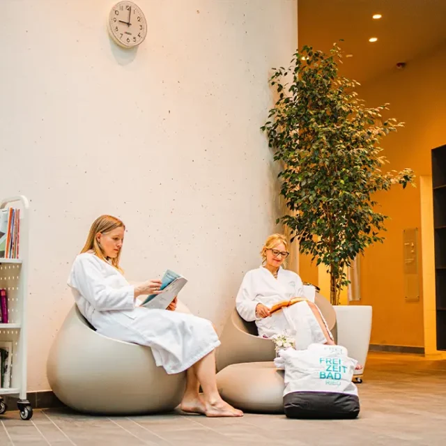 Zwei Damen im Bademantel sitzen im Ruhebereich mit Bibliothekswagen und lesen